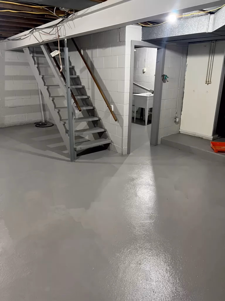 Clean basement with newly painted shiny gray floor, stairs, utility sink, and exposed ceiling beams.