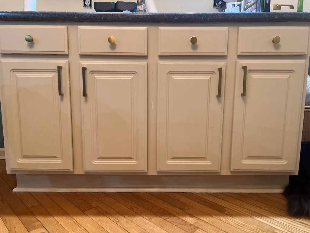 Beige kitchen cabinet with four doors and four drawers, each with different handles, on a wood floor.
