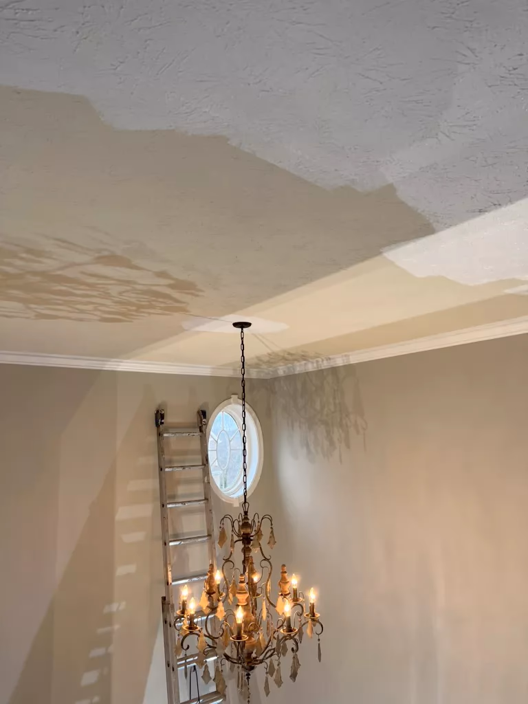 Ornate chandelier with lit bulbs hanging from a high ceiling near an oval window and a ladder.