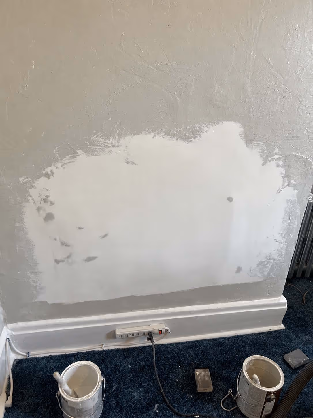 Wall section with fresh drywall patch surrounded by textured beige paint, with painting supplies on blue carpet floor.