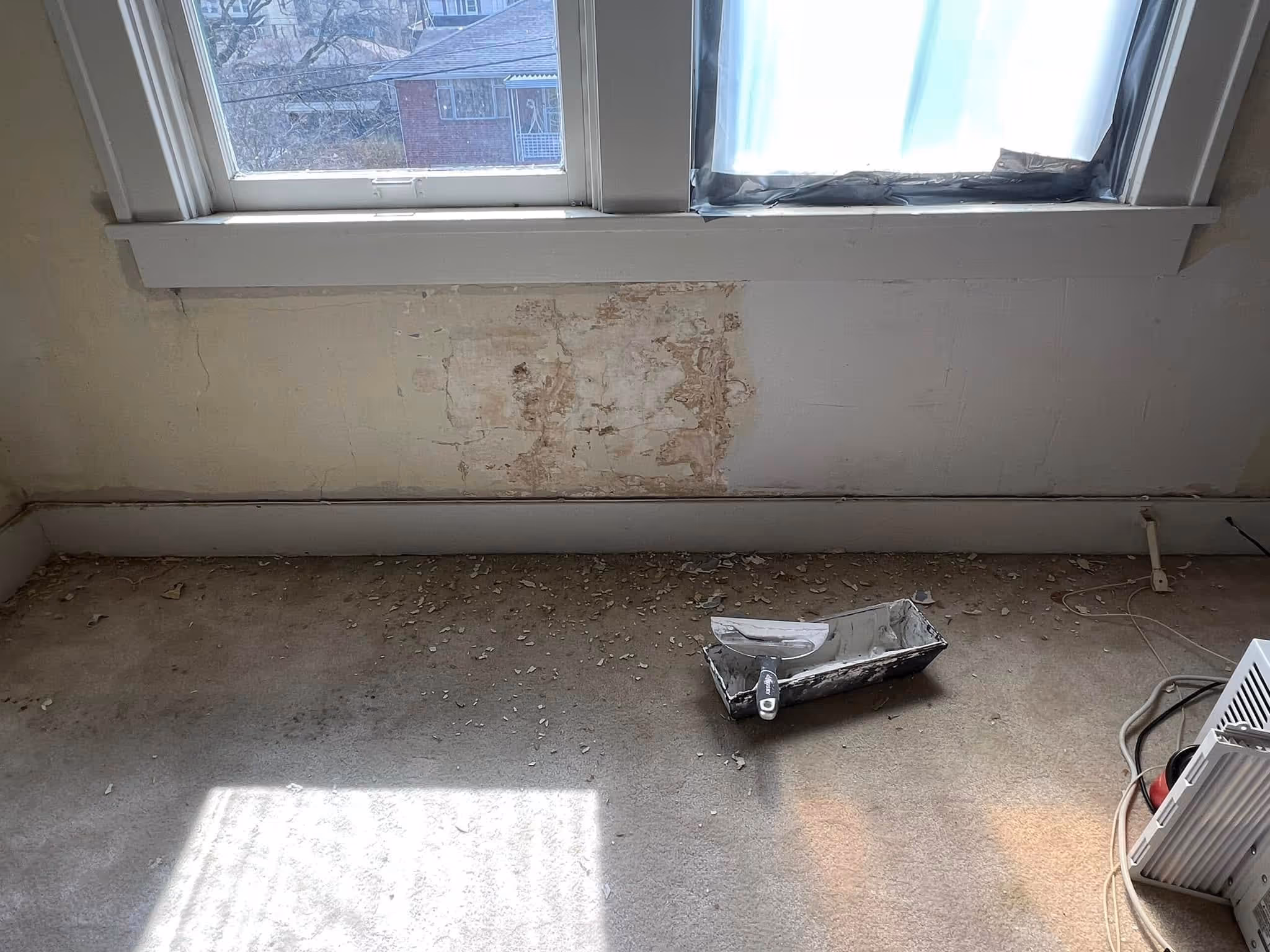 Room corner with damaged wall under two windows and a putty knife on a metal pan on the floor.