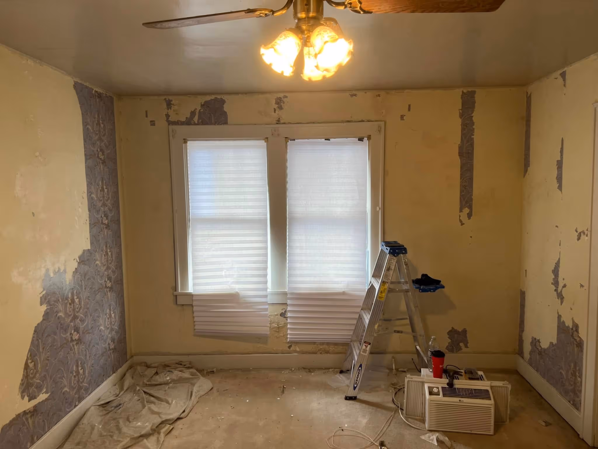 Room under renovation with peeling wallpaper, covered floor, stepladder, and window air conditioner.