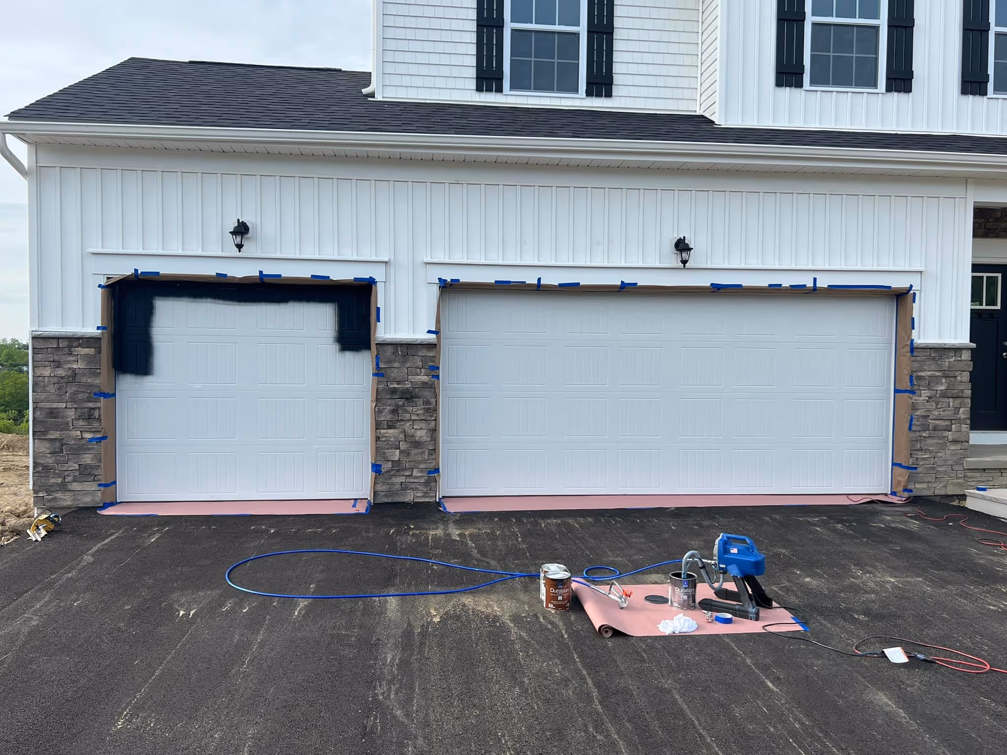 White two-car garage with the left door partially spray painted black, paint supplies on driveway.