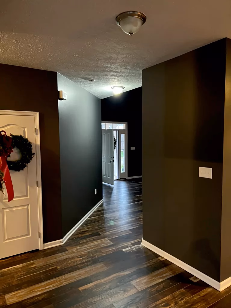 Hallway with dark walls, wood flooring, ceiling lights, and a white door decorated with a wreath and red ribbon.