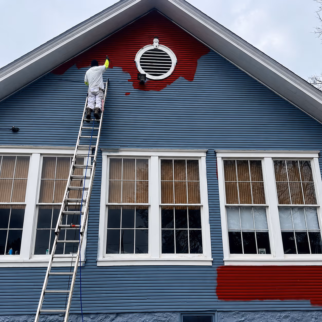 Person painting a house's blue exterior red using a ladder near a round vent under the roof.