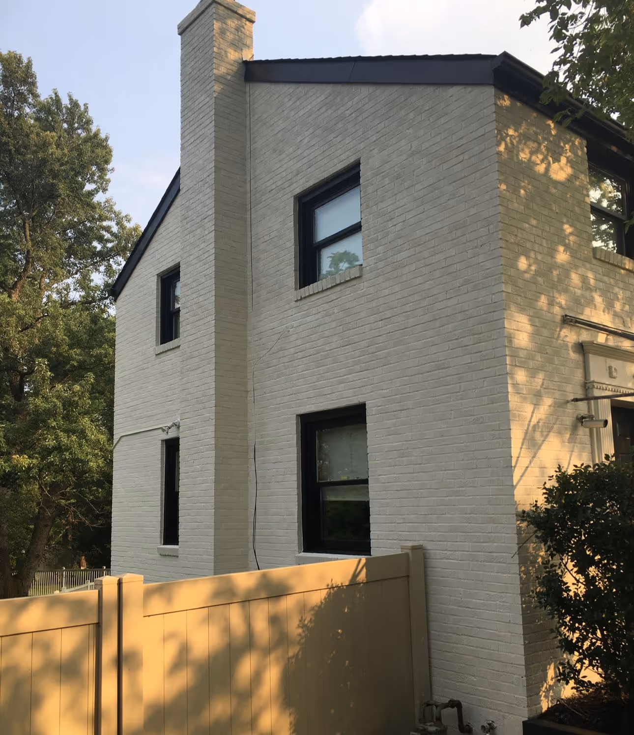Two-story beige brick house with black-trimmed windows and a tall chimney behind a light wooden fence.