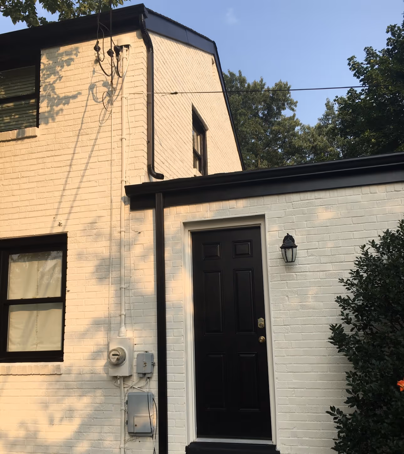 White brick house exterior with a black door, window, outdoor light, and some greenery.