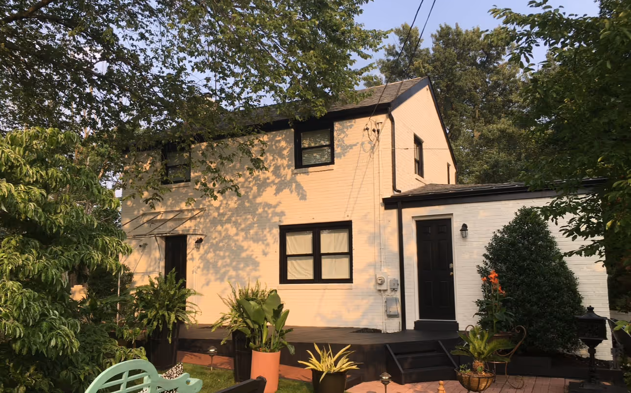 Two-story beige house with black trim surrounded by green trees and potted plants on a patio.