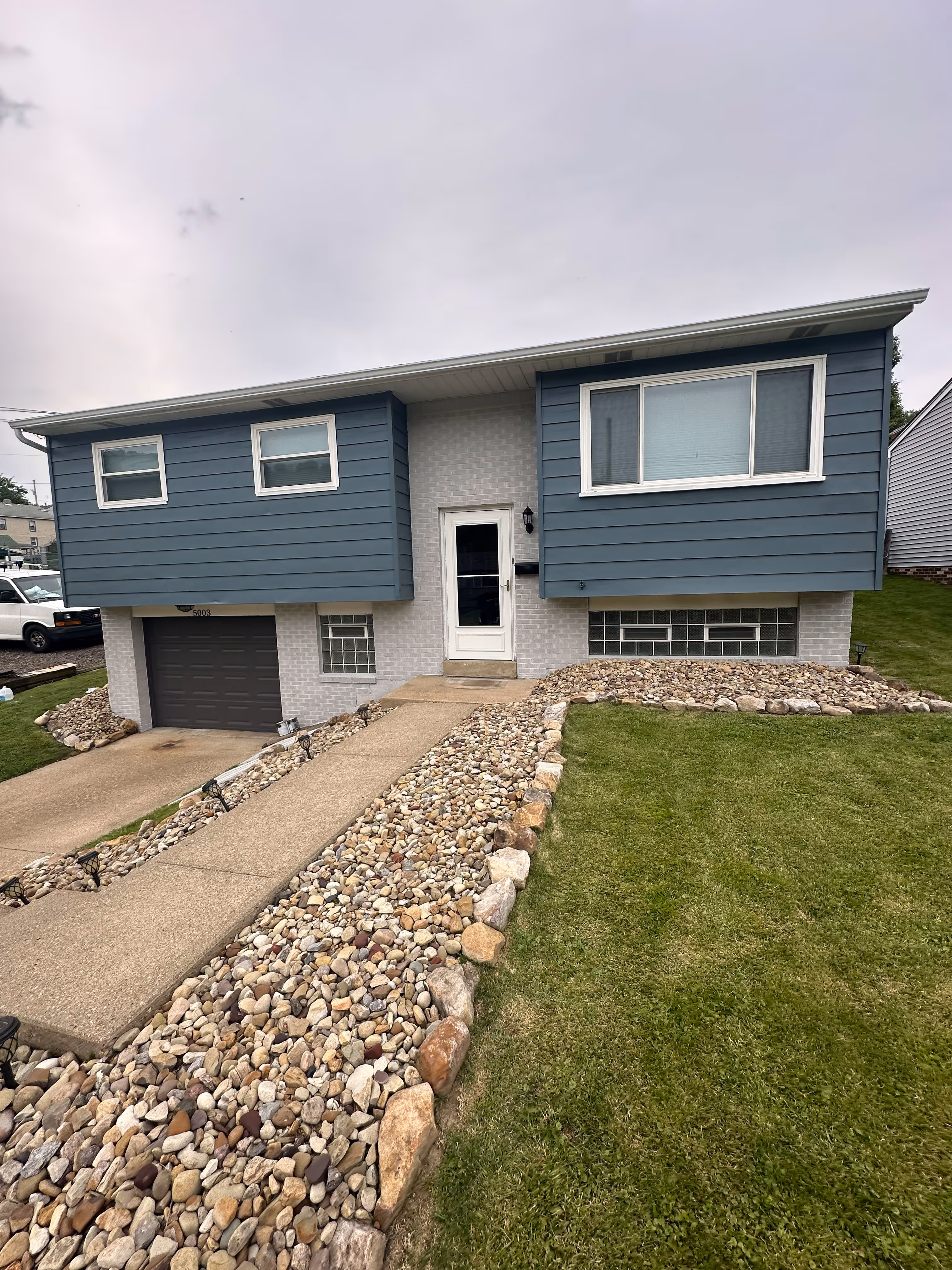 Blue and white split-level house with a concrete driveway and lawn bordered by decorative stones.