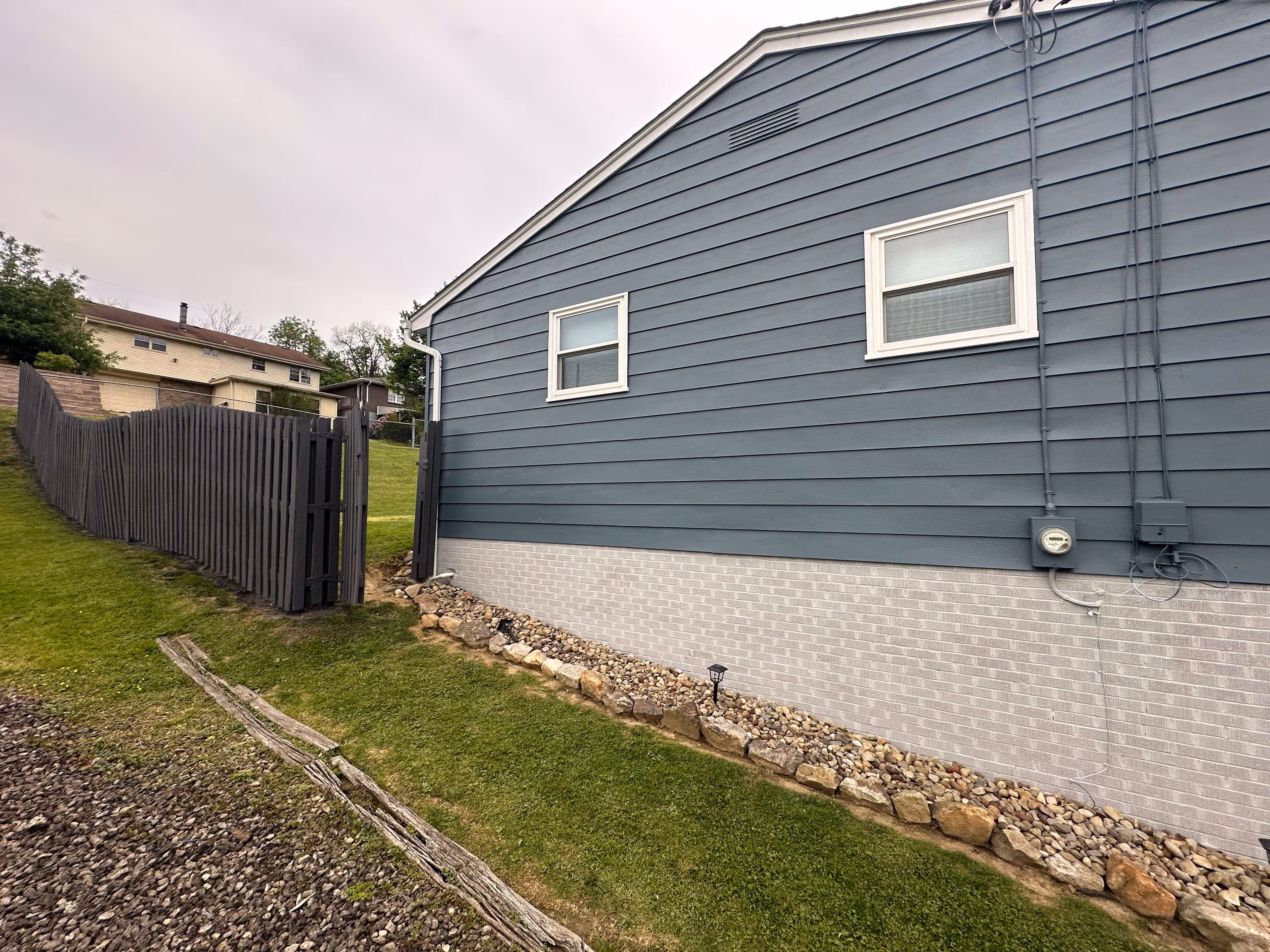 Side of a house with blue siding, white-trim windows, stone border, and adjacent tall wooden fence.