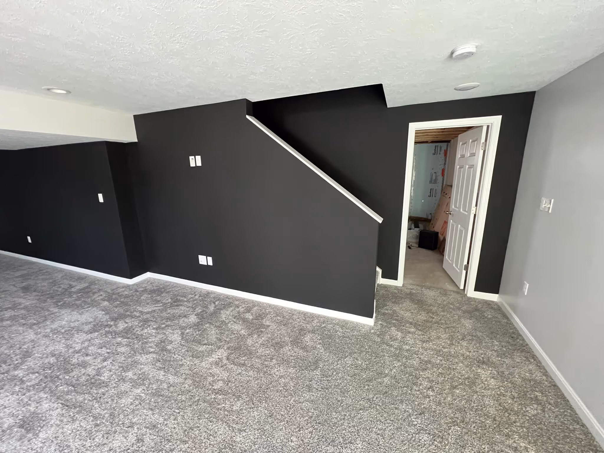 Empty room with gray carpet, black and gray walls, and an open doorway showing an unfinished space.