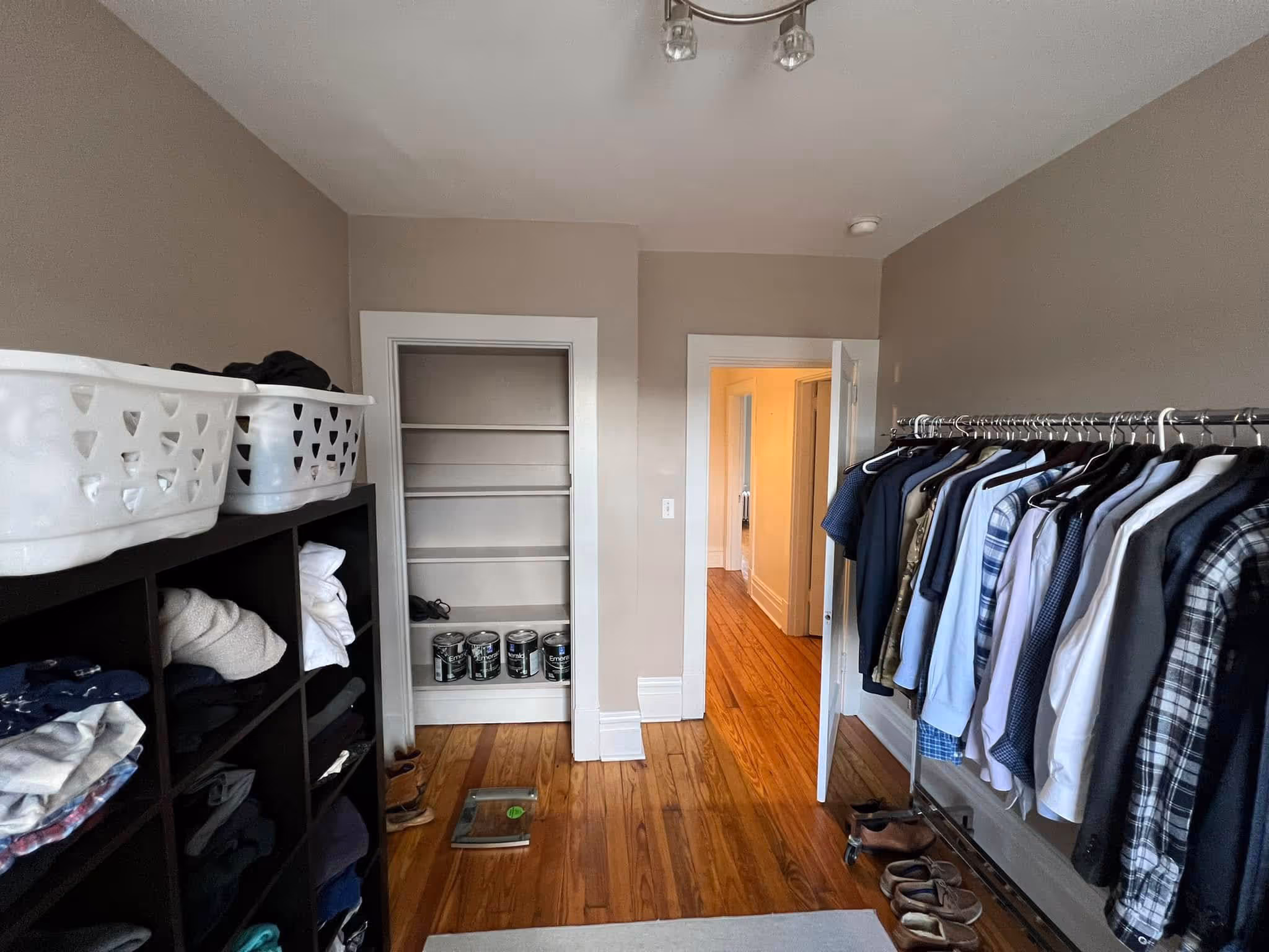 Room with wooden floor, clothes on black shelving unit and rack, open door showing hallway, and paint cans on shelves.