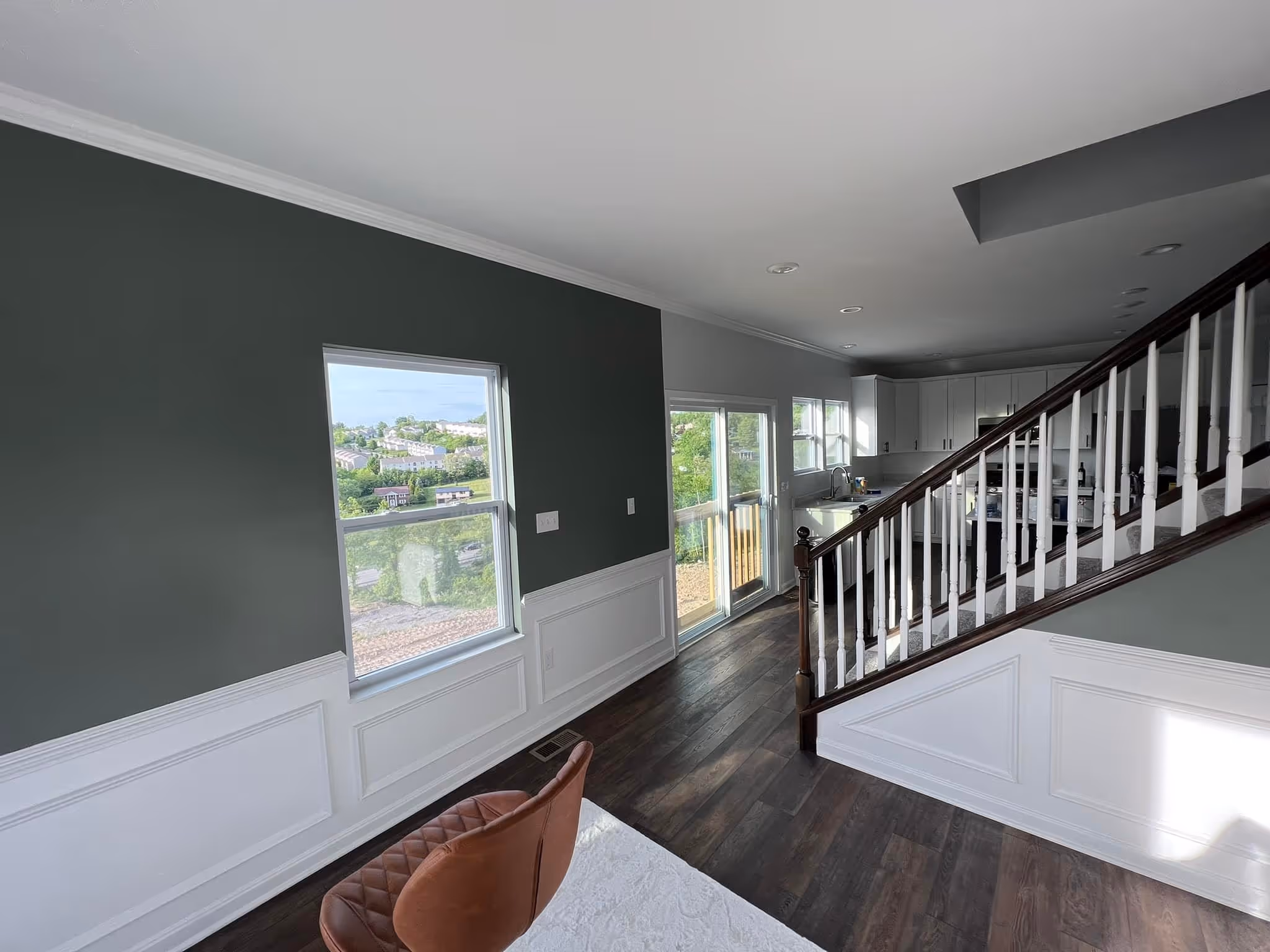 Modern open living space with dark wood flooring, white kitchen cabinets, staircase, and large windows.