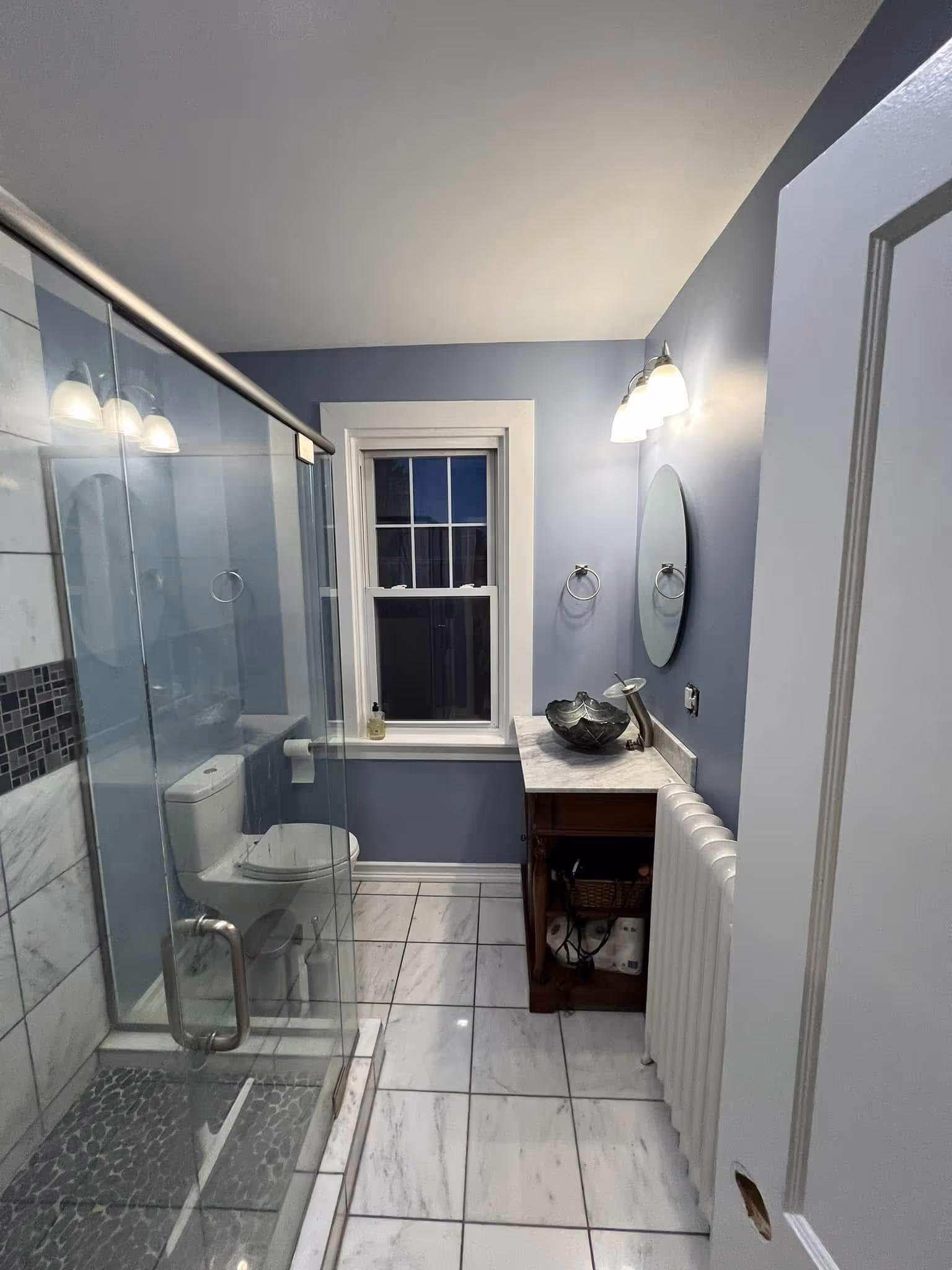 Modern bathroom with glass shower, white toilet, marble floor, blue walls, and a vanity with a vessel sink and round mirror.