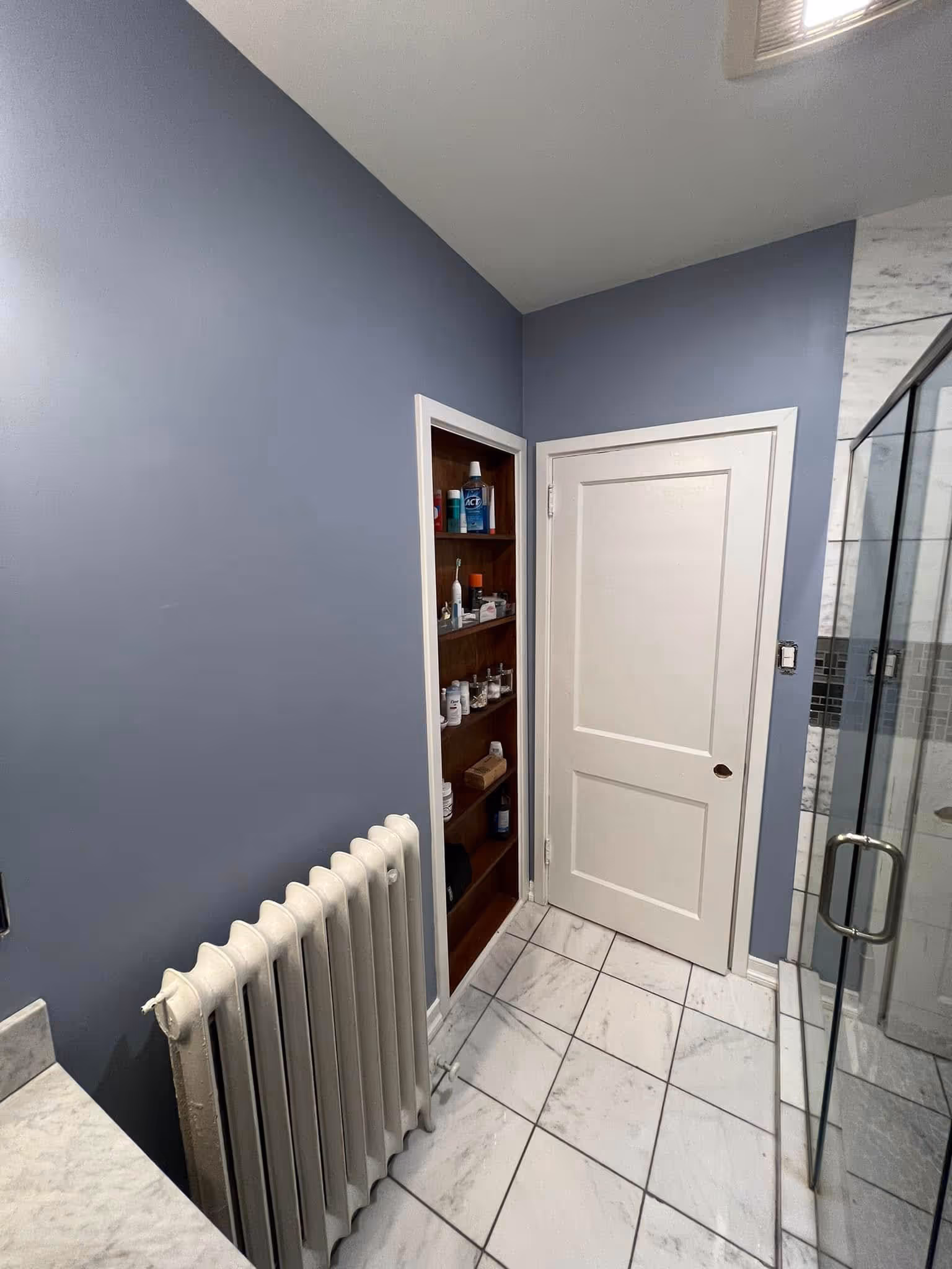 Bathroom corner with blue walls, white door, built-in wooden shelves, white radiator, and glass shower door.