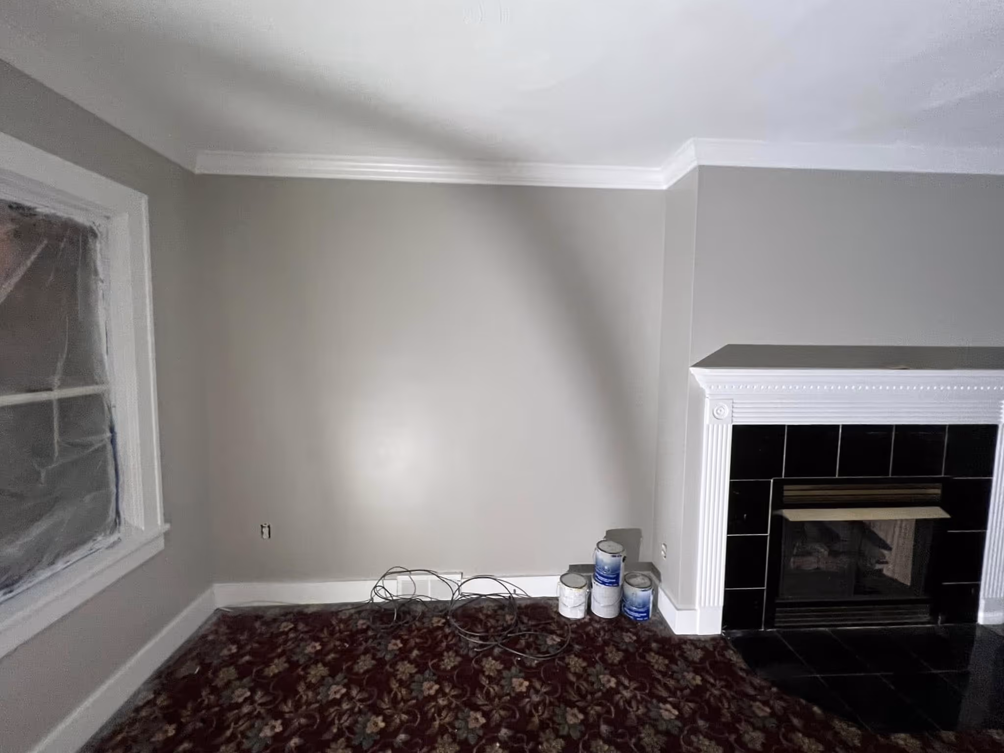 Room corner with gray painted walls, floral carpet, fireplace with black tiles, and paint cans on the floor.