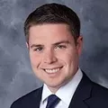 Young man with dark hair wearing a dark suit and tie, smiling against a gray background.