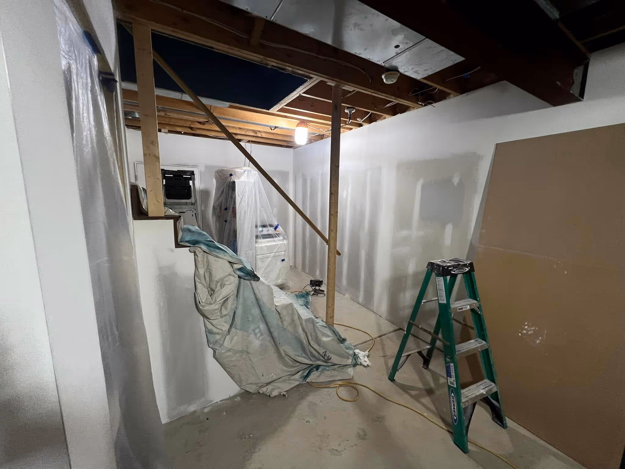 Room under renovation with exposed ceiling, covered appliances, green ladder, and plastic sheet on floor.