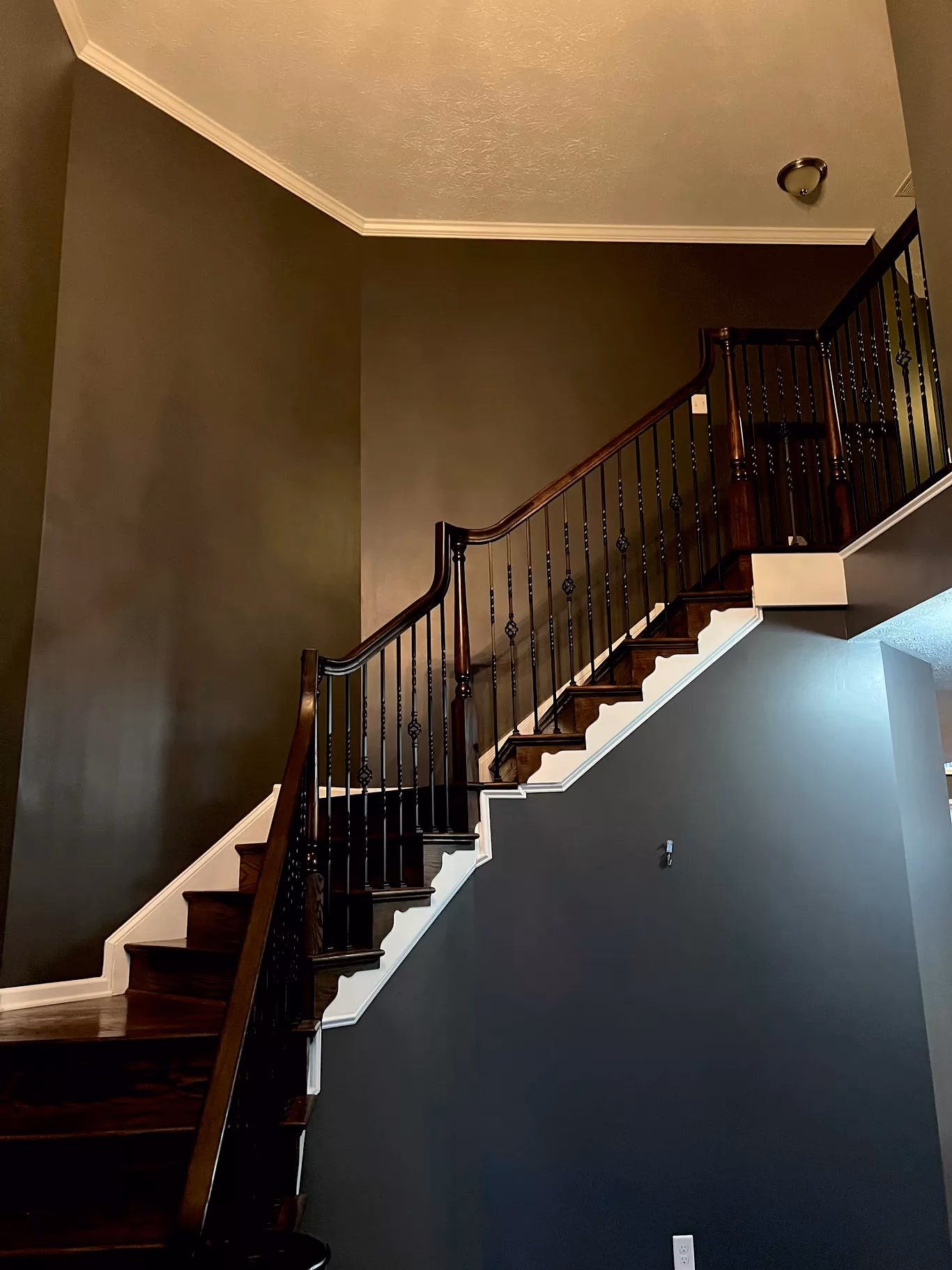 Interior view of a room with wooden floor, white staircase with dark brown railing, and an adjacent kitchen.